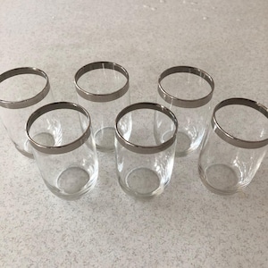 May include: A set of six clear glass tumblers, each featuring a smooth, reflective silver-toned metallic rim around the top edge. The glasses are cylindrical in shape with a simple, transparent design, arranged in a group on a flat surface.