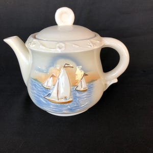 May include: A white ceramic teapot with a lid and a handle. The teapot has a blue and white design of sailboats on the sea. The teapot is decorated with gold accents.