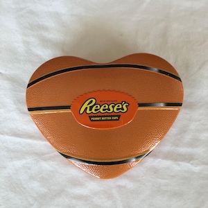 May include: A heart-shaped metal tin with a textured orange surface resembling a basketball, featuring black lines and a central orange oval label with the text Reese's Milk Chocolate Peanut Butter Cups printed in yellow and black lettering.