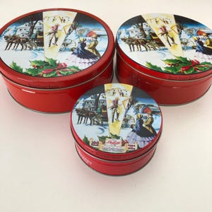 May include: Three red, round tins with lids featuring a winter scene with horse-drawn carriages and figures in period clothing. The lids have a colorful illustration with text that reads "Collin Street Bakery" and "Deluxe".
