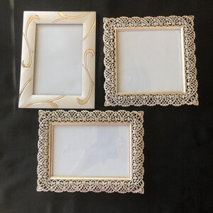 May include: Three decorative picture frames. One is rectangular with a white finish and gold swirl accents. The other two are rectangular and square with a gold filigree design. All frames have a white interior.