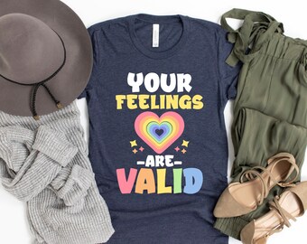 Op de afbeelding: Een marineblauw t-shirt met de tekst "Your Feelings Are Valid" in regenboogkleuren. De tekst wordt omringd door een regenboog hart en sterren.