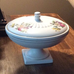 May include: White ceramic pedestal bowl with a lid. The lid has a floral design with pink, yellow, and purple flowers. The bowl has a gold trim around the top.