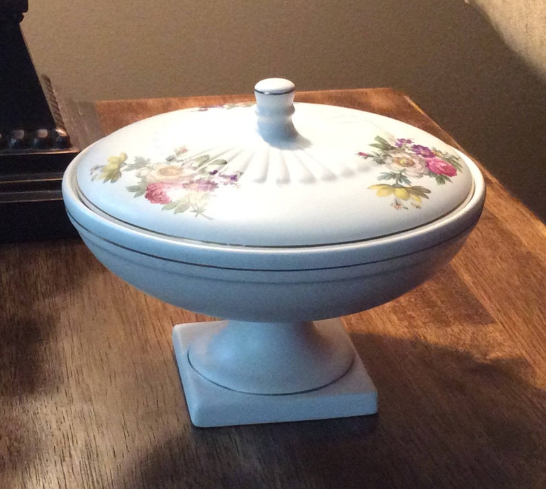 May include: White ceramic pedestal bowl with a lid. The lid has a floral design with pink, yellow, and purple flowers. The bowl has a gold trim around the top.