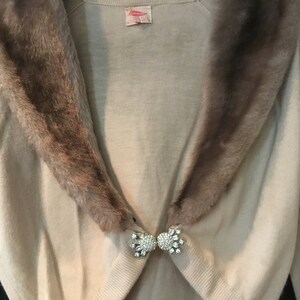 May include: A beige cardigan with a brown fur collar and a rhinestone brooch. The cardigan has long sleeves and a V-neck.