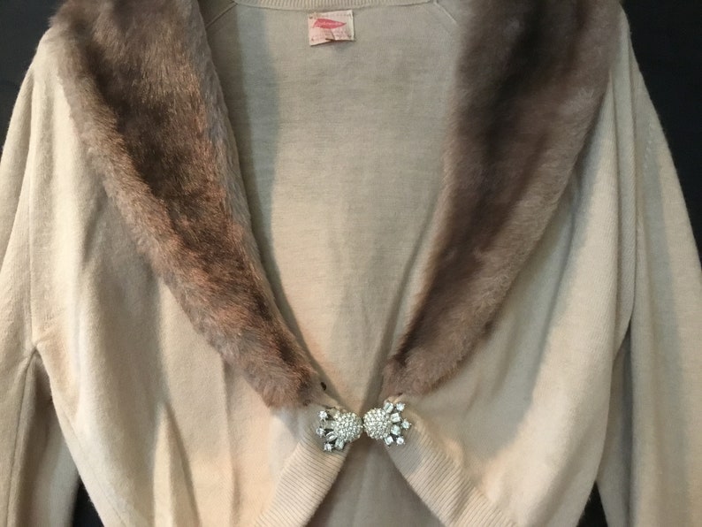 May include: A beige cardigan with a brown fur collar and a rhinestone brooch. The cardigan has long sleeves and a V-neck.