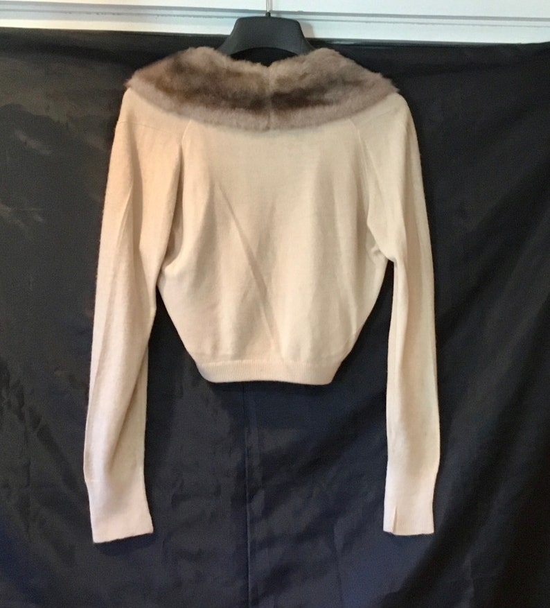 May include: A cream-colored knitted sweater with a brown fur collar. The sweater has long sleeves and a cropped length.