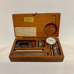 May include: A wooden box containing a dial test indicator set. The set includes a dial gauge, a clamp, a rod, and a small metal plate. The box is labeled "Wells Mfg. Co. Quality Tools Compton, Calif."