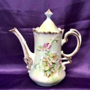 May include: A white porcelain teapot with a floral design. The teapot has a gold trim and a lid with a finial. The flowers are pink, white, and green.