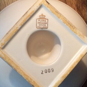May include: White ceramic base with a square shape and a round hole in the center. The base has a gold inscription that reads "Flora Keramiek Gouda Holland" and the number "2009" printed on the bottom.