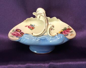 May include: A blue and white porcelain serving dish with a gold trim and floral design. The dish has a handle and a pedestal base.