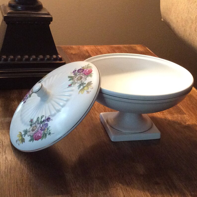 May include: White ceramic pedestal bowl with a floral design on the lid. The bowl has a gold trim around the top and bottom.