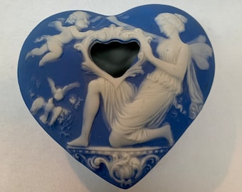 May include: A blue and white ceramic heart-shaped trinket box with a raised design of a woman and a cherub. The box has a small opening in the center.