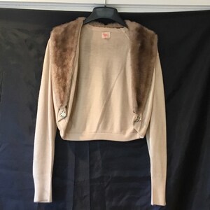 May include: A beige knitted cardigan with a brown faux fur collar. The cardigan has two decorative buttons on the front.