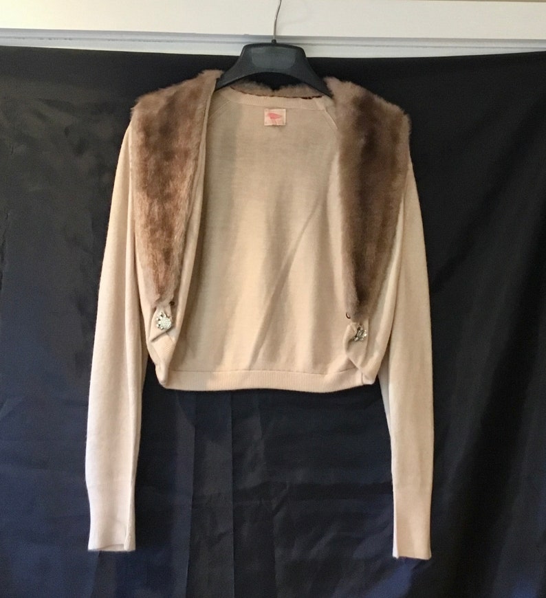 May include: A beige knitted cardigan with a brown faux fur collar. The cardigan has two decorative buttons on the front.