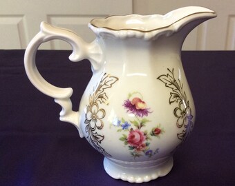 May include: A white porcelain creamer with a gold trim and floral design. The creamer has a scalloped rim and a handle.