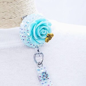 May include: A badge reel featuring a light blue rose with a small yellow bee accent. The reel has a clear, glittery base and a silver clip. The badge reel is designed to hold an ID badge or other small items.