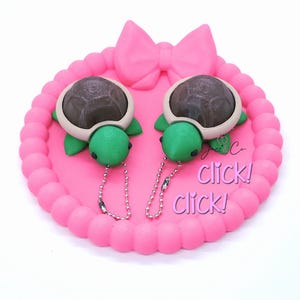 May include: Two turtle keychains with brown shells, green bodies, and silver chains rest on a pink, bow-shaped tray. The tray has a beaded edge and the words "Click! Click!" are written on the tray.