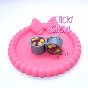May include: A pink, bow-shaped tray with a beaded edge holds two miniature gray pots filled with colorful food. The tray has the words "Click! Click!" in pink script. The pots have silver handles and are filled with what appears to be a stew.