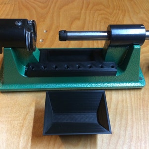 May include: A green reloading press with a black handle and a black tray with multiple holes. A black tray is in the foreground.