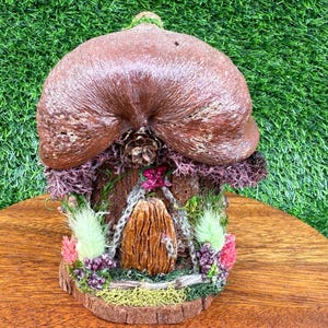 Puede incluir: Una casa de hadas en miniatura con un techo en forma de hongo marrón y una puerta de madera. La casa está decorada con musgo, flores y vegetación, y se encuentra sobre una superficie de madera sobre un fondo verde.
