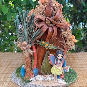 May include: A handcrafted fairy house with a thatched roof, featuring a miniature fairy figurine. The house is made of natural materials, including wood, dried leaves, and moss. The fairy has blue and yellow wings and a yellow skirt.