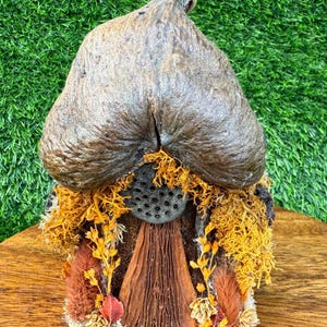 Puede incluir: Una casa de hadas artesanal con un gran techo marrón texturizado. La casa presenta una puerta de madera, una ventana circular y está adornada con musgo y follaje naranja y amarillo. La base es de madera.
