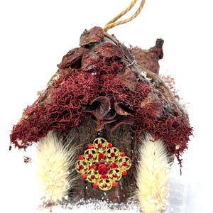 Puede incluir: Adorno decorativo en miniatura con forma de casa rústica. El tejado está cubierto de musgo rojo y corteza, con un adorno dorado y rojo en la parte delantera. Tiene un lazo de cuerda natural para colgar y detalles en crema.