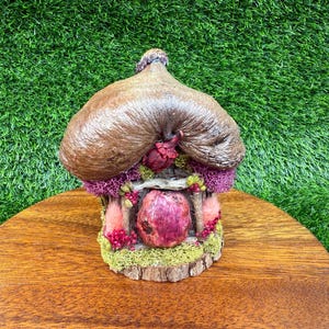 Pode incluir: Uma figura de casa de fadas caprichosa com um telhado de colmo castanho e uma porta rosa. A casa é adornada com elementos decorativos roxos, rosa e verdes, e assenta sobre uma base de madeira. A miniatura está sobre um fundo verde.