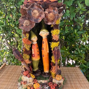 May include: A whimsical autumn-themed diorama featuring two gnome figures in orange and green attire. The scene includes miniature pumpkins, dried flowers, and decorative moss, all set within a rustic, bark-like frame. The overall design evokes a fairytale forest setting.