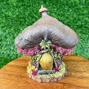Puede incluir: Una casa de hadas en miniatura con un tejado de paja marrón y una puerta amarilla. La base es de madera y está rodeada de flores y musgo de colores. La casa tiene un aspecto caprichoso y artesanal.
