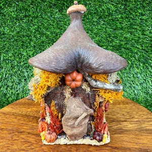 以下が含まれることがあります： 茶色のキノコ型の屋根と小さなカボチャのアクセントが付いた、手作りの妖精の家。家は、オレンジと黄色の葉とミニチュアのかぼちゃに囲まれた、質感のある樹皮のようなファサードが特徴です。
