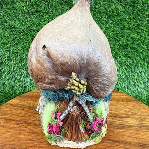 May include: A whimsical, handcrafted fairy house sculpture. The house features a bulbous, textured brown top with a gold accent. The base is adorned with miniature flowers, moss, and a small wooden structure, creating a charming, naturalistic aesthetic.