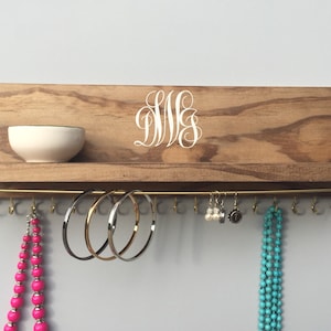 May include: Wooden jewelry organizer with a shelf and hooks. The organizer has a light brown finish, a white monogram, and a brass bar with hooks for hanging jewelry. A small white bowl sits on the shelf, and various necklaces and bracelets are displayed.
