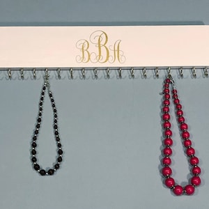May include: White wooden jewelry organizer with the monogram "BBH" in gold lettering. The organizer has 16 hooks and two necklaces hanging from it. One necklace is black and the other is pink.