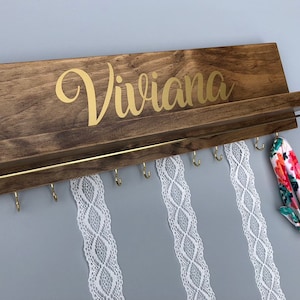 May include: Wooden wall-mounted headband organizer with the name "Viviana" in gold lettering. It has a shelf and hooks for storing headbands and hair accessories. There are three white lace headbands hanging from the hooks and a floral headband on the shelf.