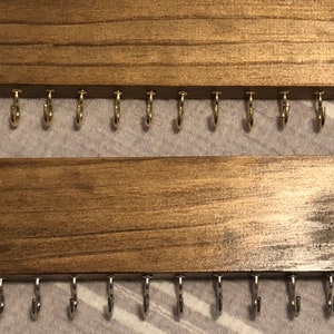 Puede incluir: Dos organizadores de joyas de pared de madera con ganchos. La fila superior tiene ganchos dorados y la fila inferior tiene ganchos plateados.