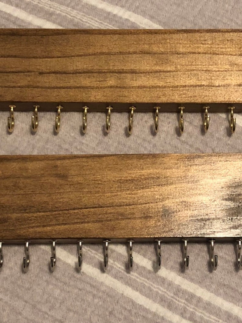 Puede incluir: Dos organizadores de joyas de pared de madera con ganchos. La fila superior tiene ganchos dorados y la fila inferior tiene ganchos plateados.