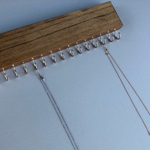 Puede incluir: Un organizador de collares de madera con 15 ganchos. Dos collares cuelgan de los ganchos. Uno es plateado y el otro es dorado.