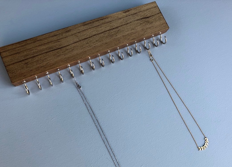 Puede incluir: Un organizador de collares de madera con 15 ganchos. Dos collares cuelgan de los ganchos. Uno es plateado y el otro es dorado.