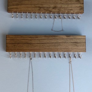 Puede incluir: Dos organizadores de joyas de madera con ganchos de metal. El organizador superior tiene un brazalete plateado colgando de &eacute;l. El organizador inferior tiene dos collares colgando de &eacute;l.