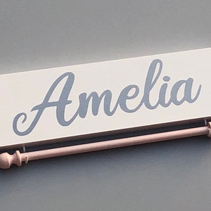 May include: A white wooden sign with the name "Amelia" written in cursive in gray paint. The sign is attached to a pink wooden dowel rod with decorative finials.