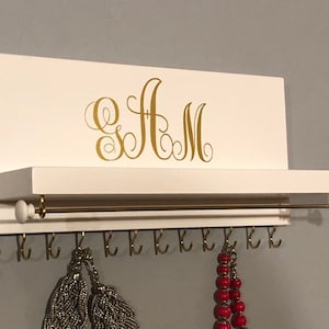 May include: White wooden jewelry organizer with a gold metal rod and hooks. The organizer has a shelf and a gold monogram "AM" on the front.