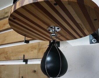 May include: A wooden speed bag platform with alternating light and dark wood stripes. A black leather speed bag hangs below, attached by a metal swivel. The platform is mounted to the wall with metal brackets, creating a boxing setup.