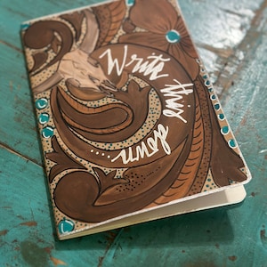 May include: A brown leather-bound journal with a hand-painted design featuring a skull, floral patterns, and the words "Write this down".