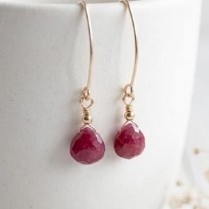 May include: A pair of gold-colored drop earrings with teardrop-shaped, faceted, deep red gemstones. The earrings hang from delicate gold wires and are displayed against a white background.