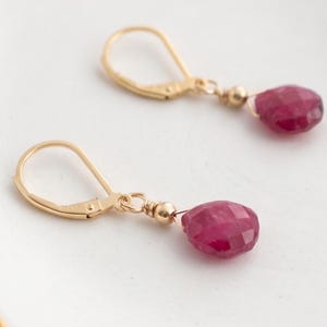 May include: A pair of gold-colored leverback earrings with faceted, teardrop-shaped ruby gemstones. The earrings are displayed on a white surface with a gold trim.