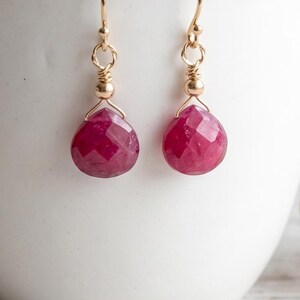 May include: A pair of gold-coloured drop earrings. Each earring features a faceted, teardrop-shaped, deep pink ruby gemstone. The earrings hang from gold-coloured ear wires and have small gold beads.