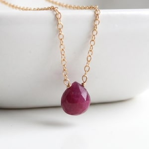 May include: A delicate gold chain necklace featuring a faceted, teardrop-shaped ruby pendant. The ruby is a deep, rich red color, and the chain is a fine, linked design. The necklace is displayed against a white background.