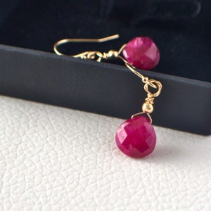 Peut inclure: Boucles d'oreilles pendantes dorées avec des pierres précieuses rouge foncé facettées. Les boucles d'oreilles ont un design simple avec une fermeture à crochet et une petite perle dorée. Les pierres sont en forme de goutte.